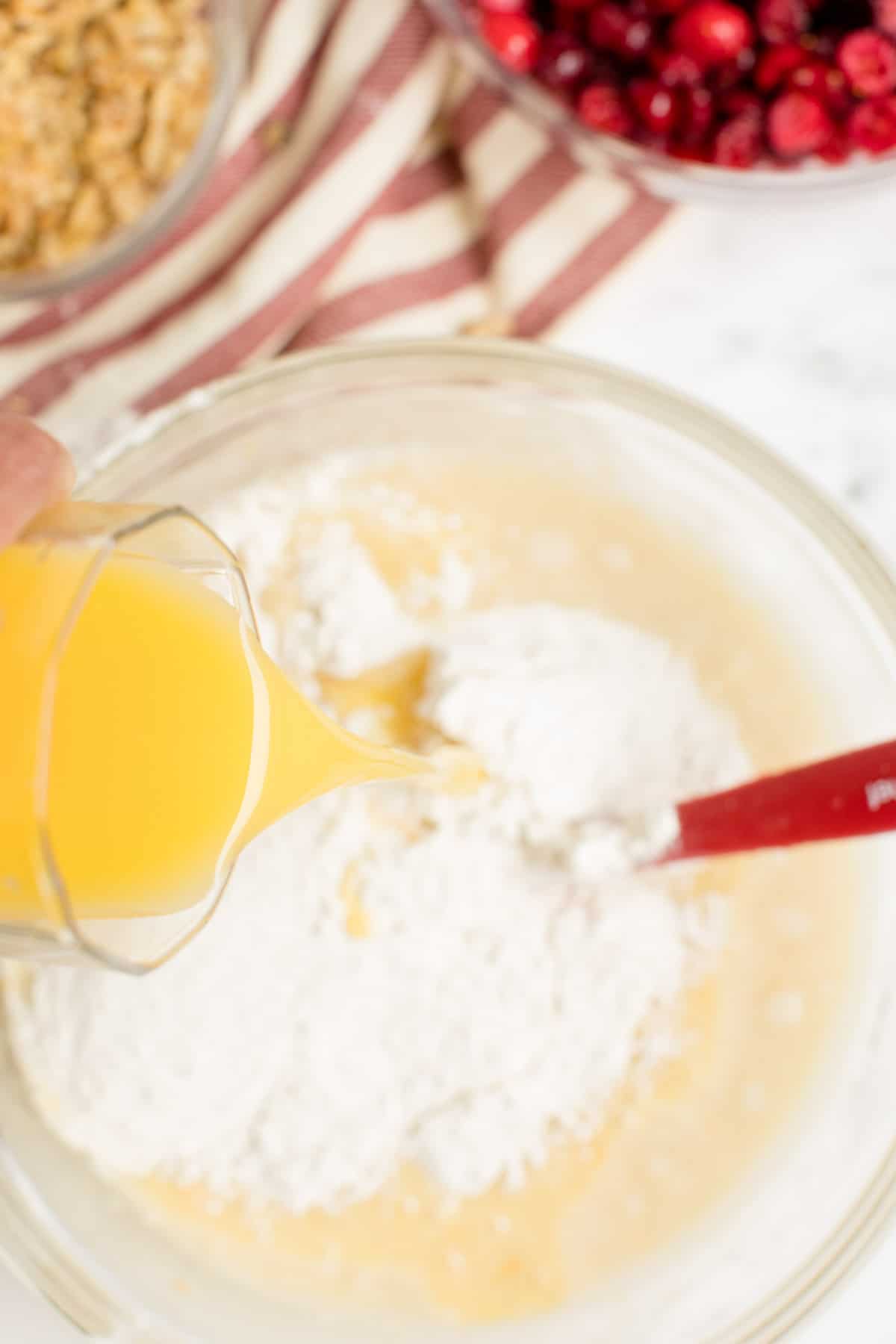 Hand pouring a glass of orange juice into a bowl with cranberry orange bread batter