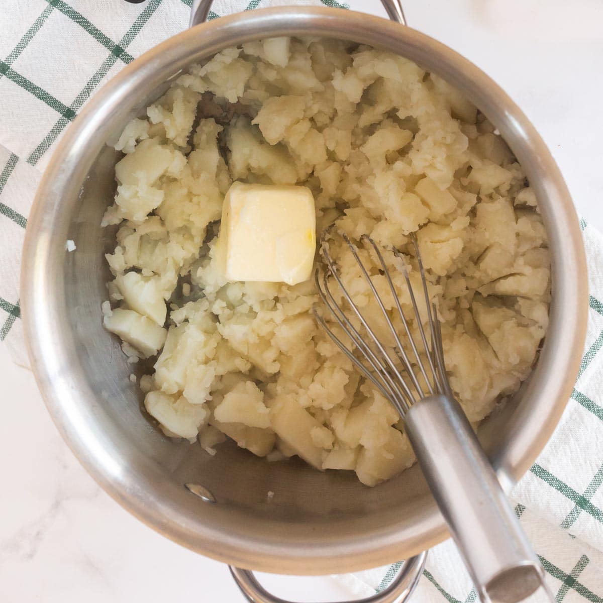 Metal pot with mashed potatoes, a metal whisk, and a cube of butter