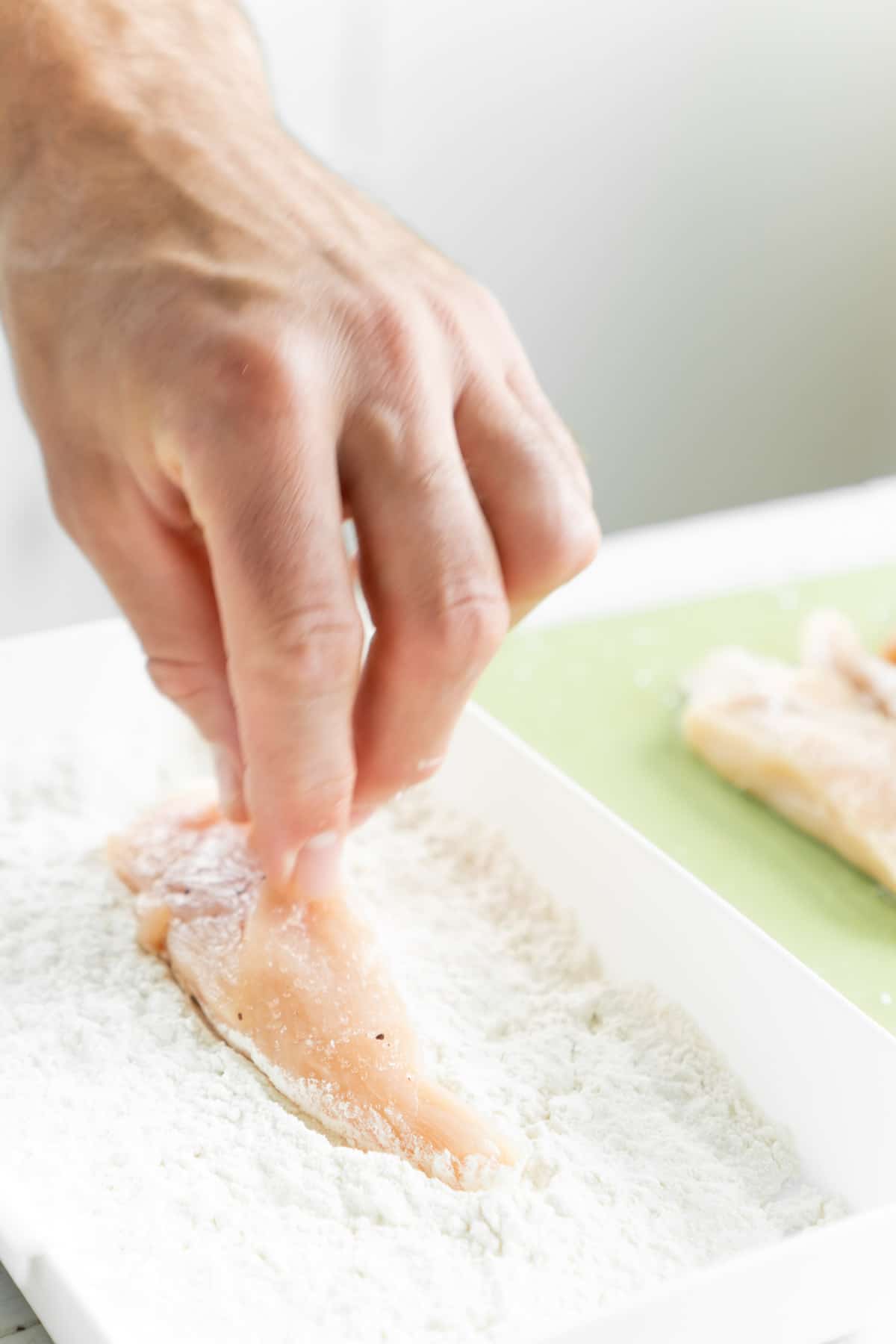 Hand putting chicken tender into dish of flour