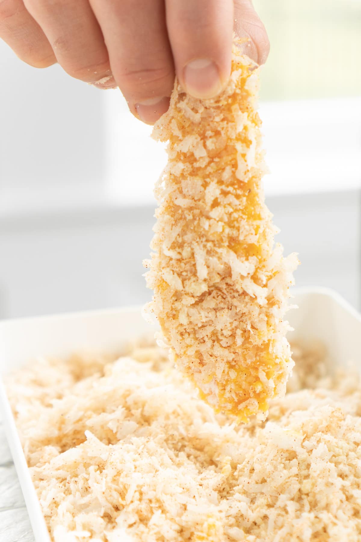 Hand holding up a breaded coconut chicken tender above coconut chicken breading