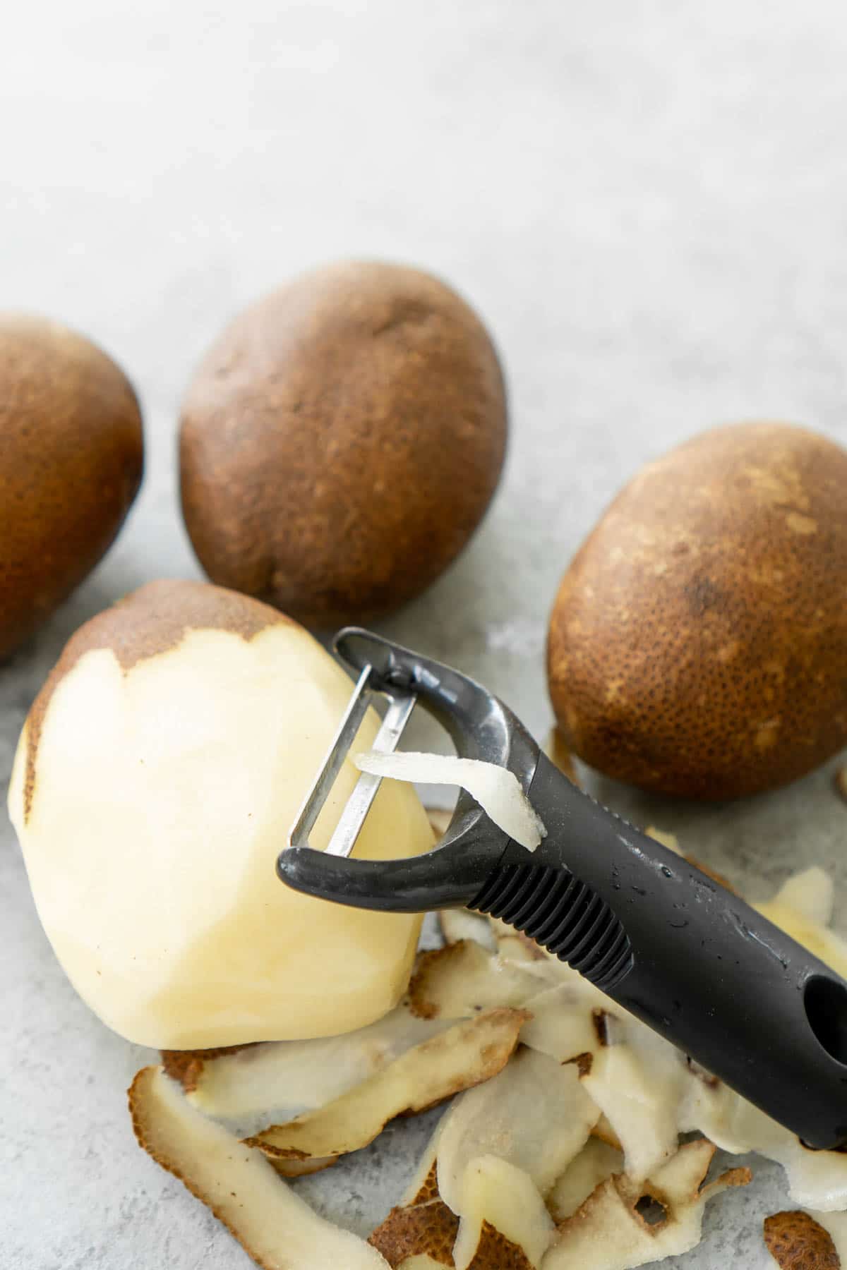 Three potatoes with a half peeled potato, potato peeler, and pile of potato peelings