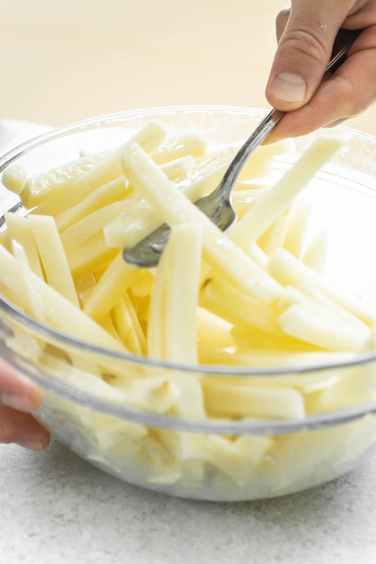 A hand holding a spoon mixing cut potatoes in a glass bowl