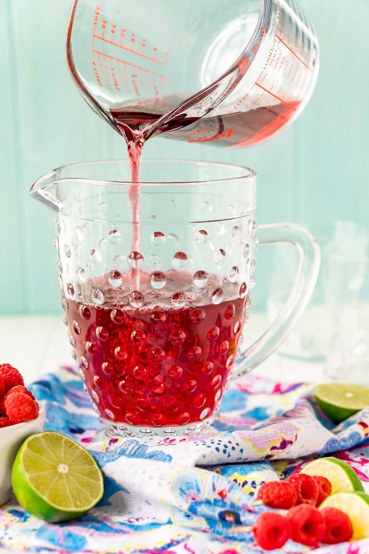 A glass measuring cup pouring raspberry juice into a glass pitcher