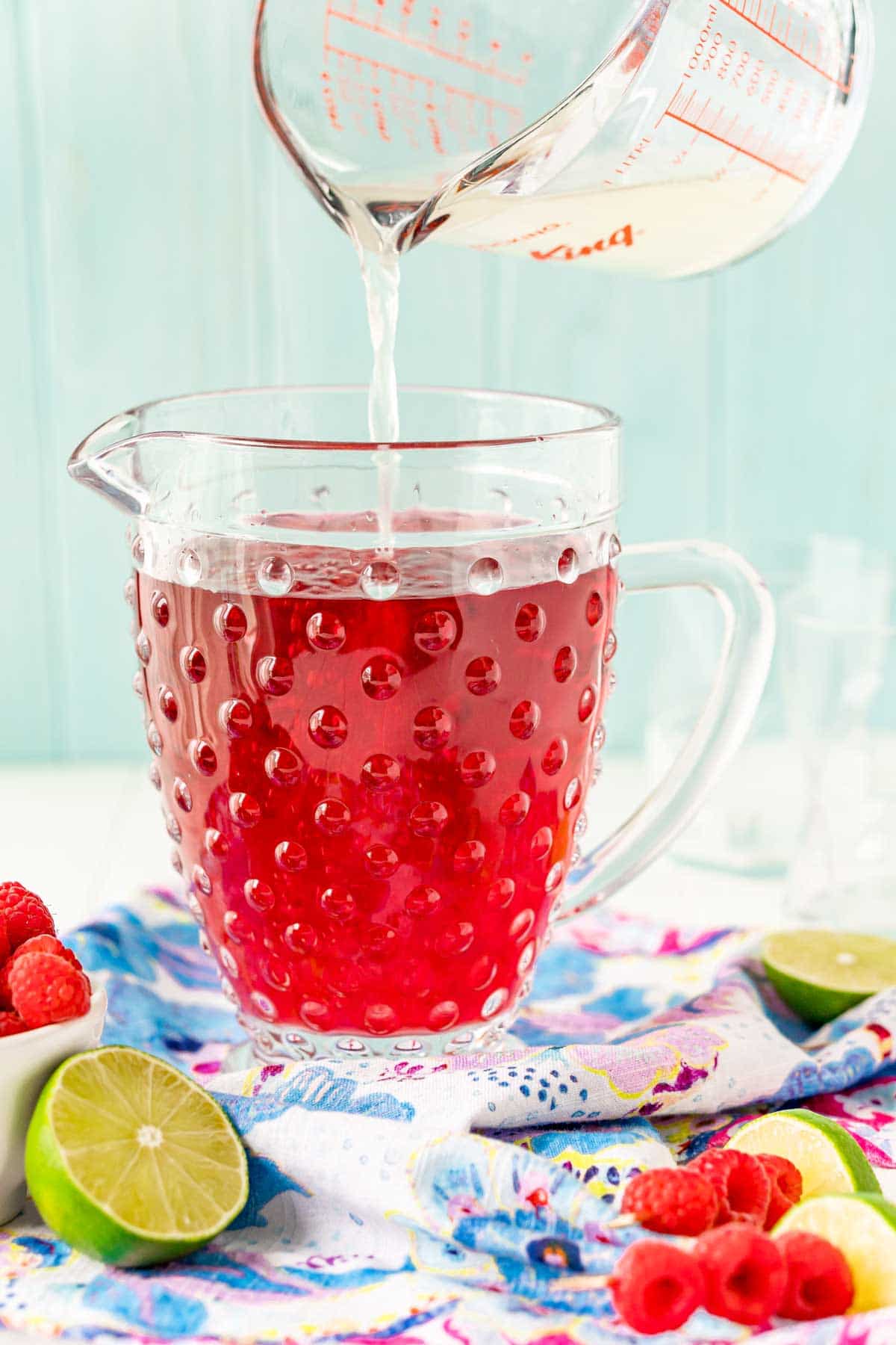 A measuring cup pouring clear liquid into a pitcher of red raspberry punch