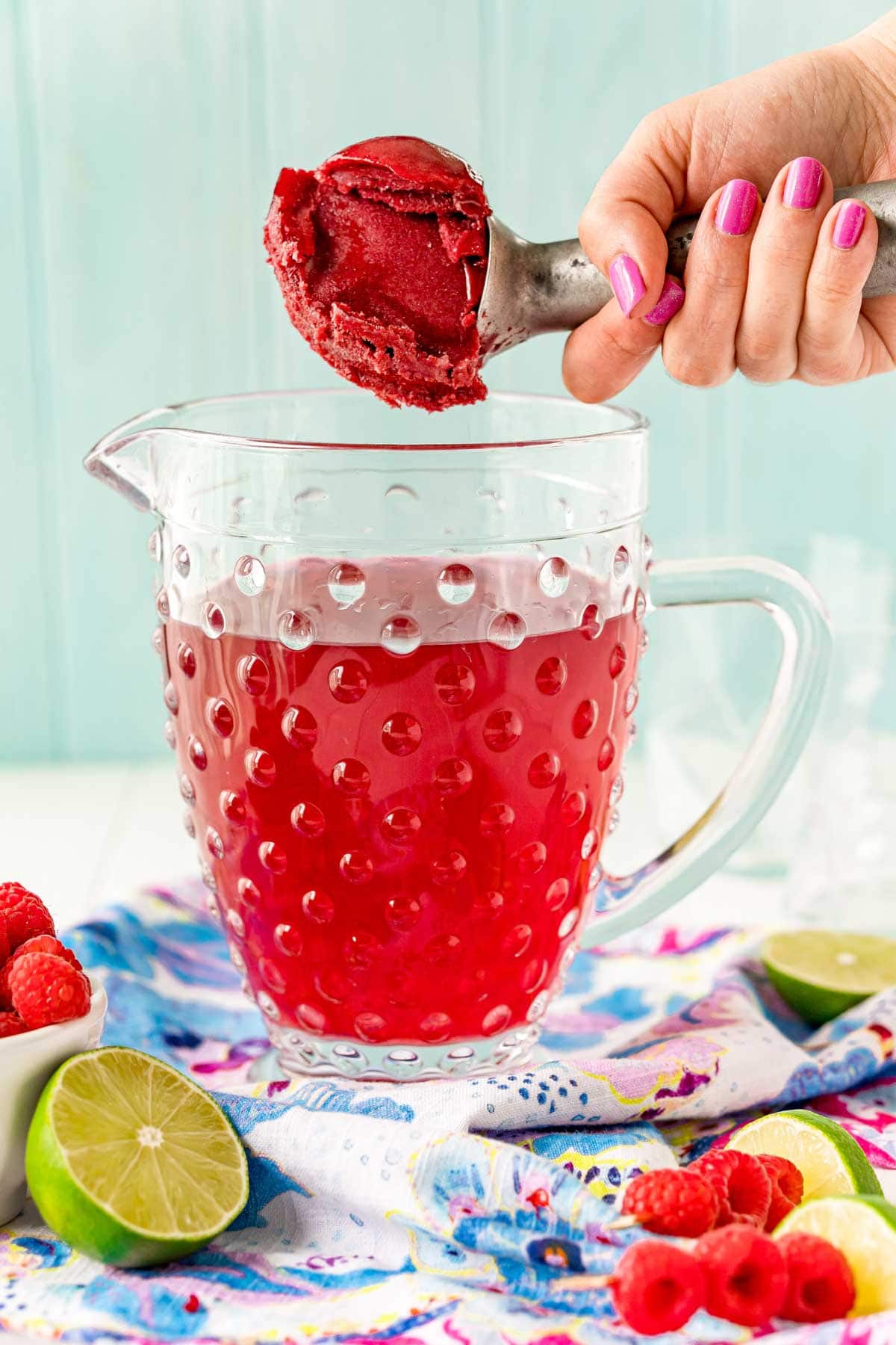 A hand holding an ice cream scoop with raspberry sorbet and a pitcher with raspberry juice
