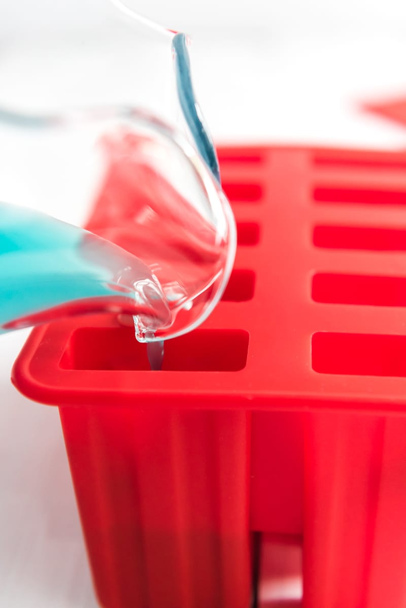 A glass pitcher pouring blue lemonade into popsicle molds