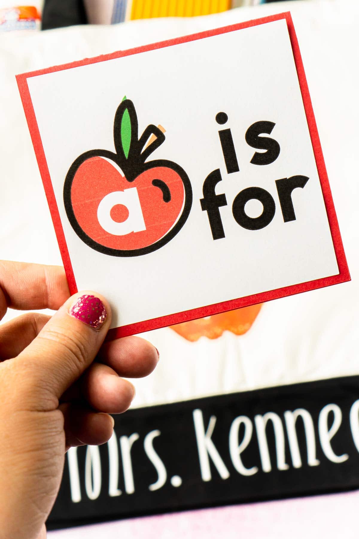 A woman's hand holding an A is for card over a tote bag