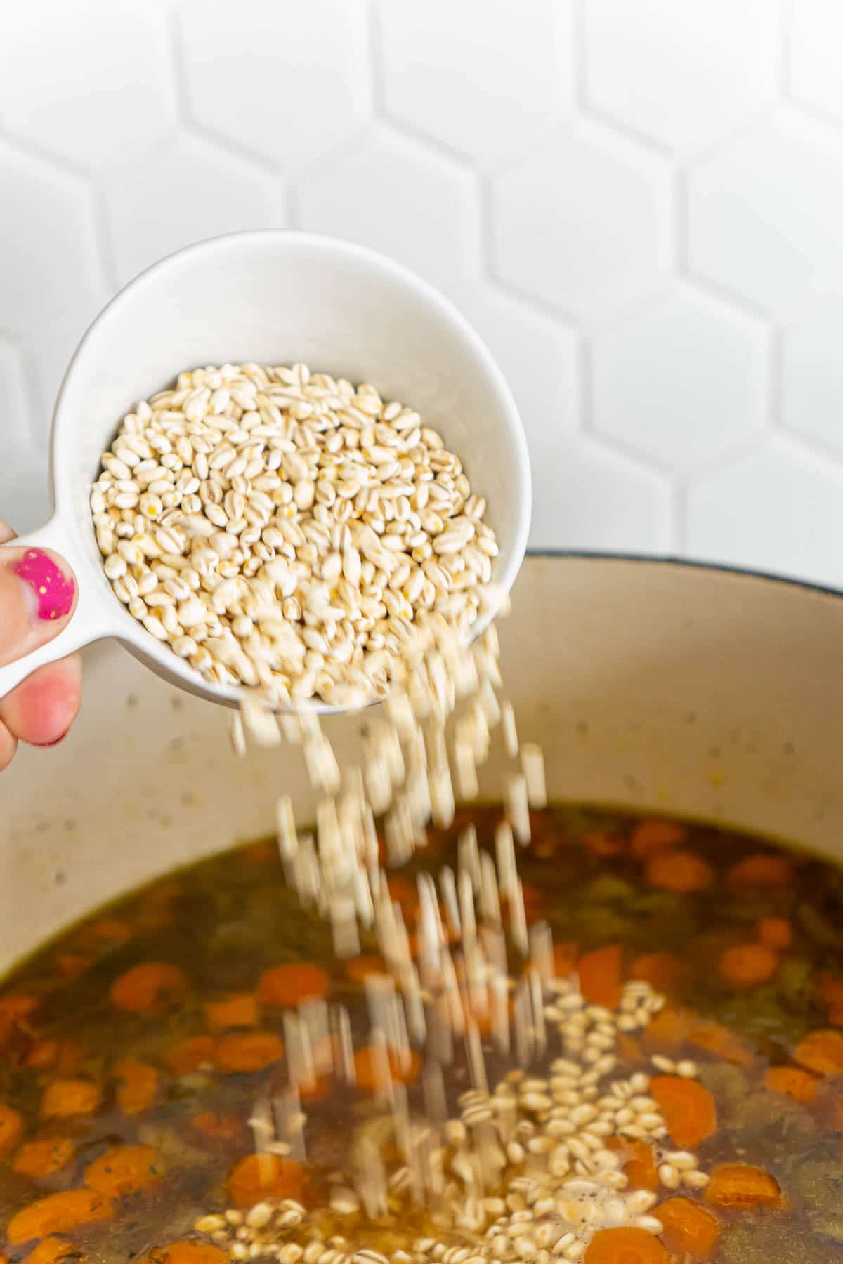 Woman's hand pouring in a cup of barley into a beef barley soup