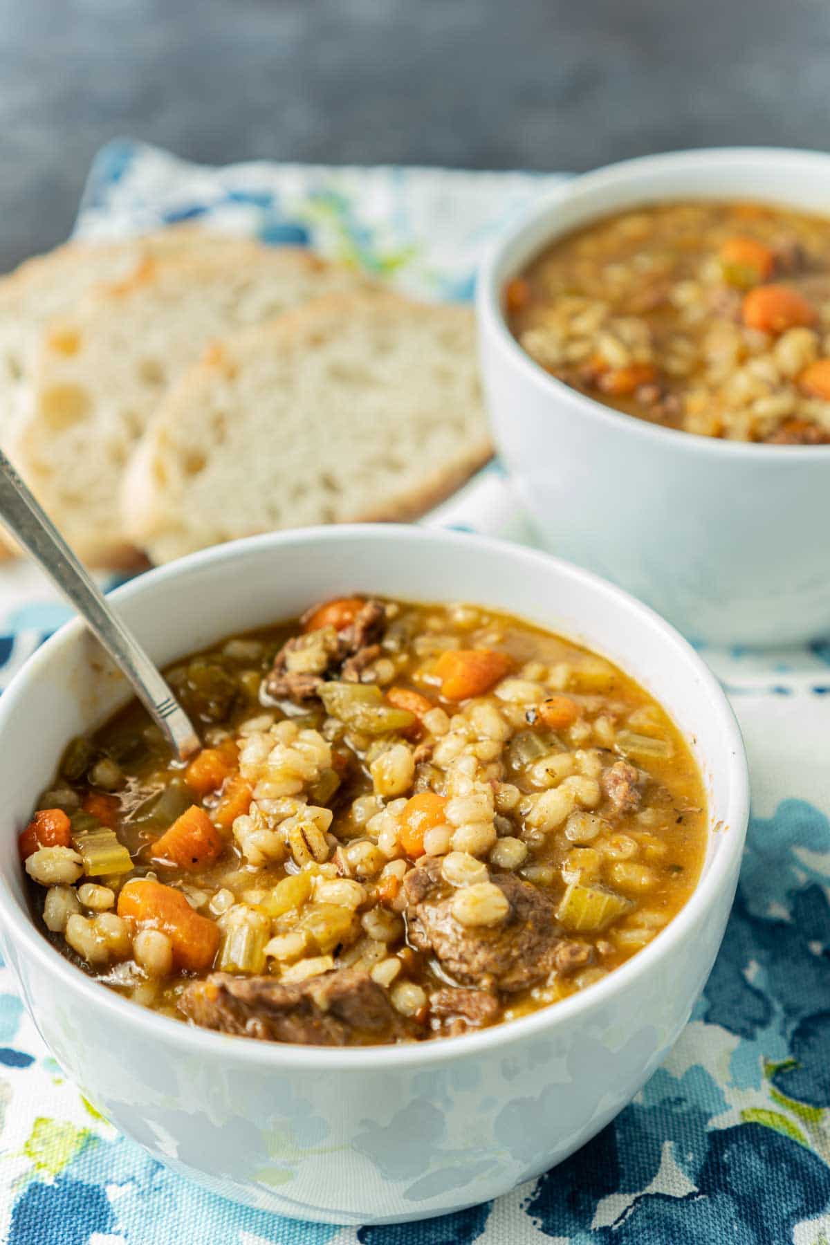 Two white bowls full of beef barley soup with bread in the background