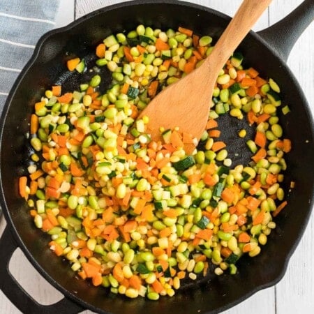 A cast iron pan with a wooden spoon and a bunch of chopped veggies for corn succotash