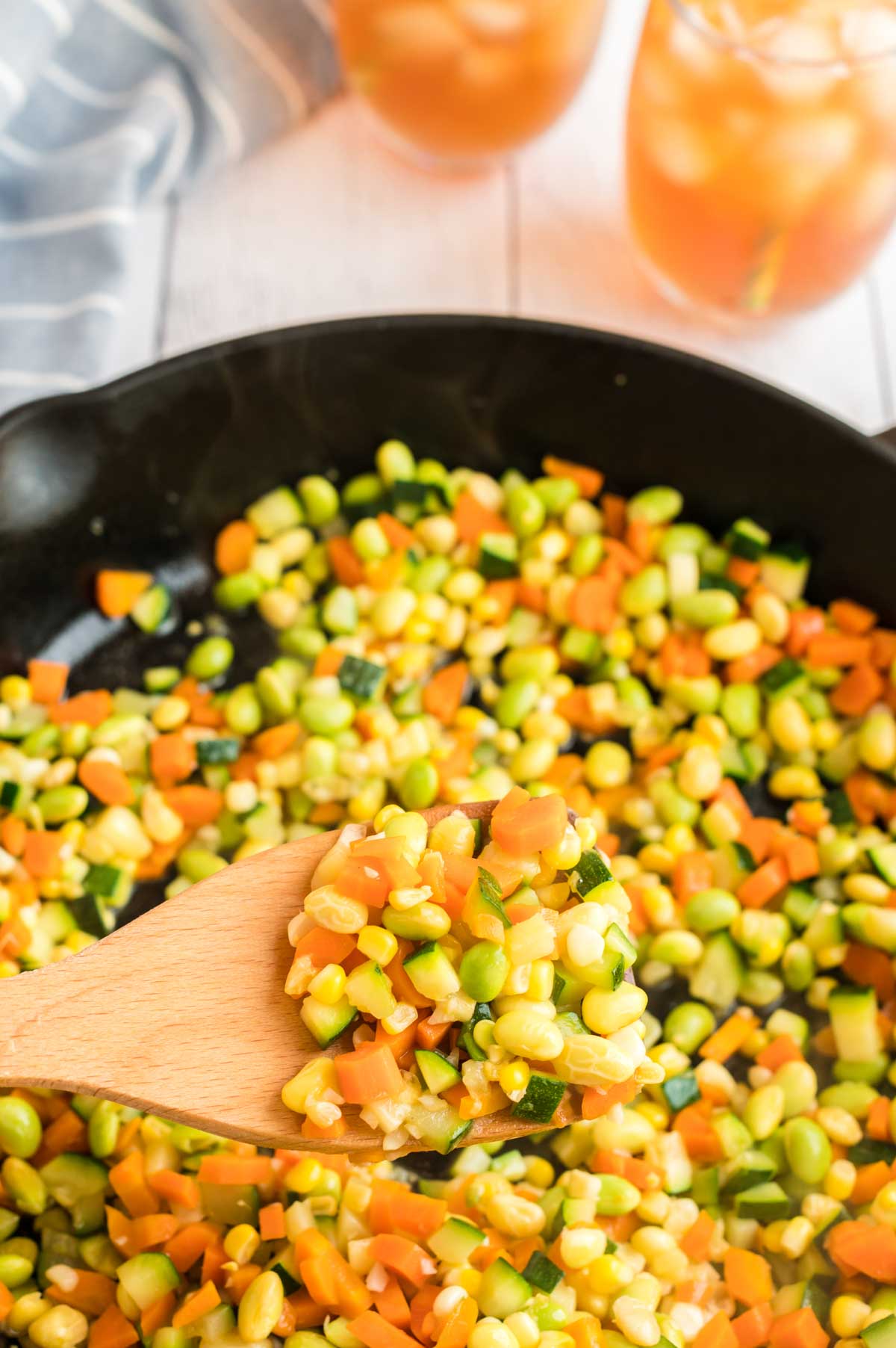 A wooden spoon with corn succotash above a cast iron pan with corn succotash