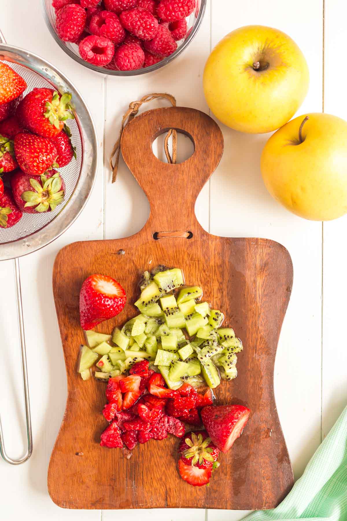 Cutting board with diced strawberries and kiwi with fruit around on the table