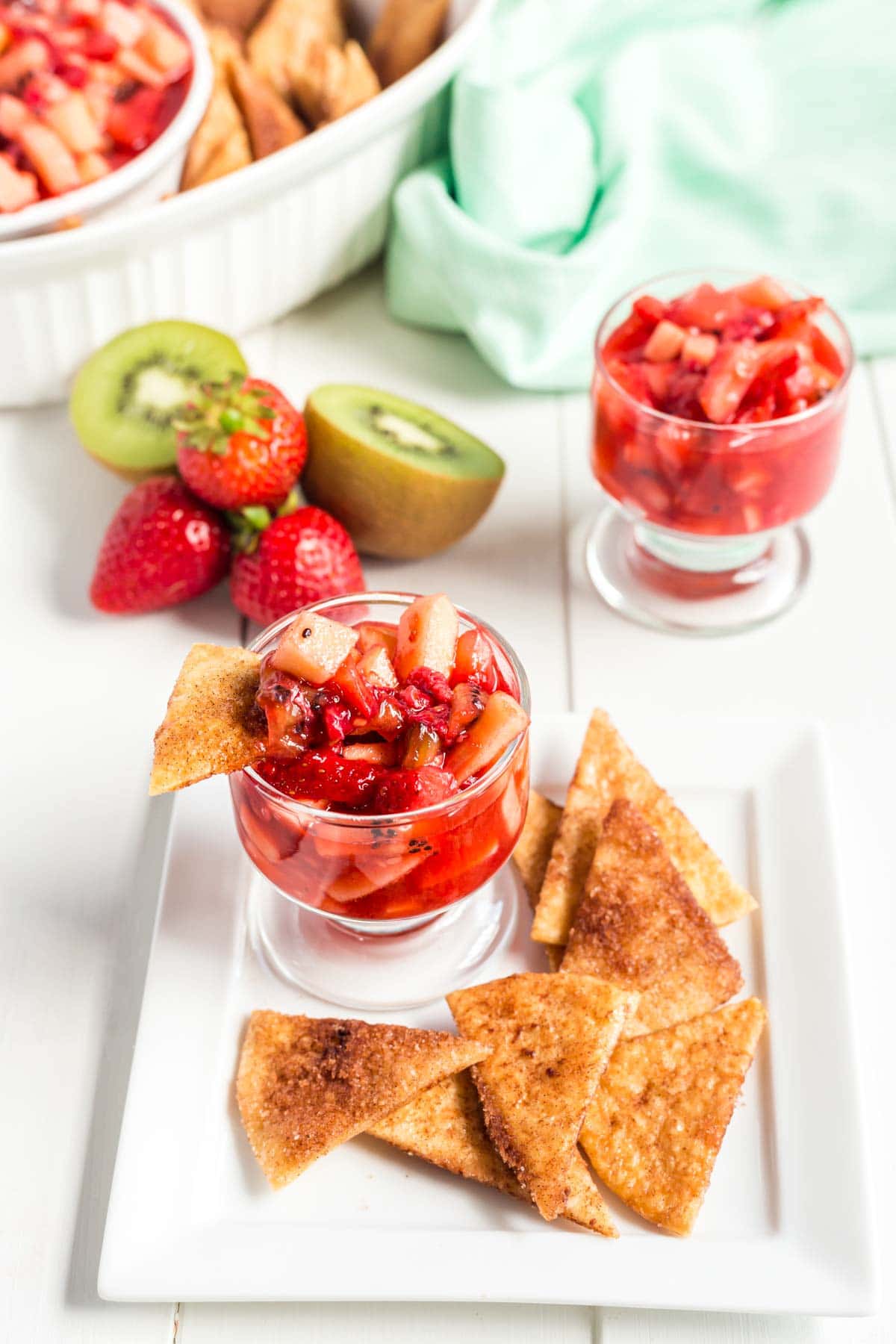 A small glass of fruit salsa on a plate with cinnamon chips and fruit salsa in the background