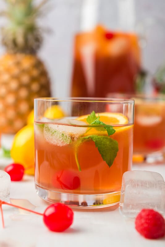 Glass of fruit punch in front of a pitcher of fruit punch with fruit all around