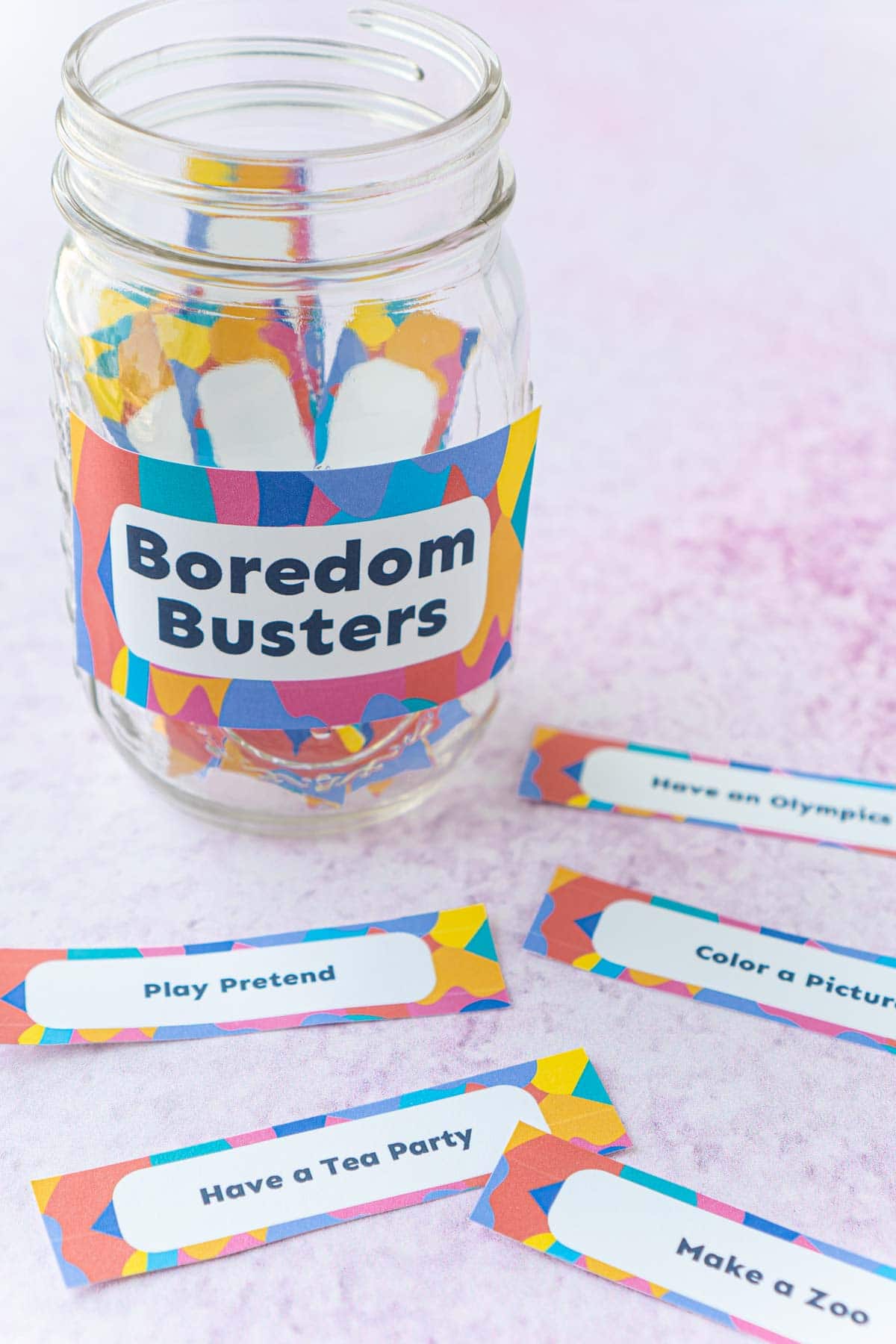 A mason jar with a boredom buster label and a bunch of boredom busters on the table in front of it