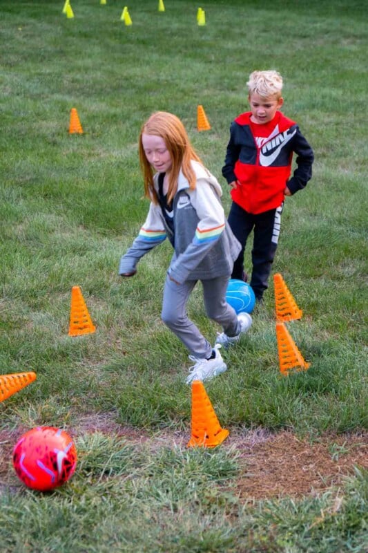 Two kids kicking soccer balls through cones