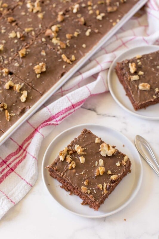 A chocolate brownie cake square on a white plate with a sheet pan of cake in the background