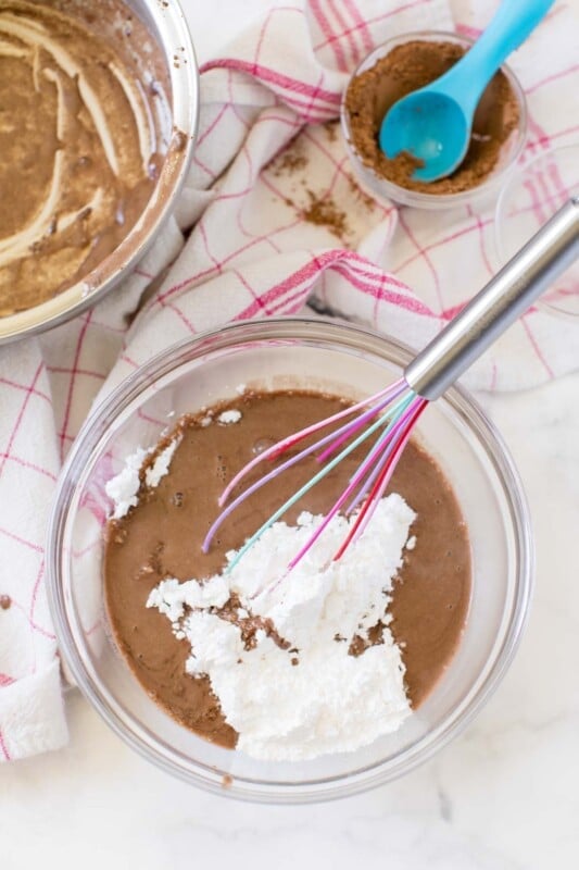 Powdered sugar and chocolate brownie cake icing in a bowl with a pink whisk