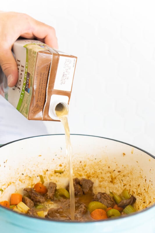 Hand pouring a container of beef broth into beef stew