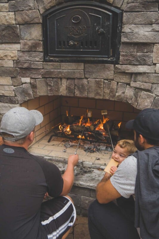 Two men and babies making s'mores in a fireplace