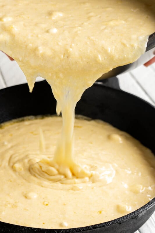 A metal bowl pouring cornbead batter into a cast iron skillet