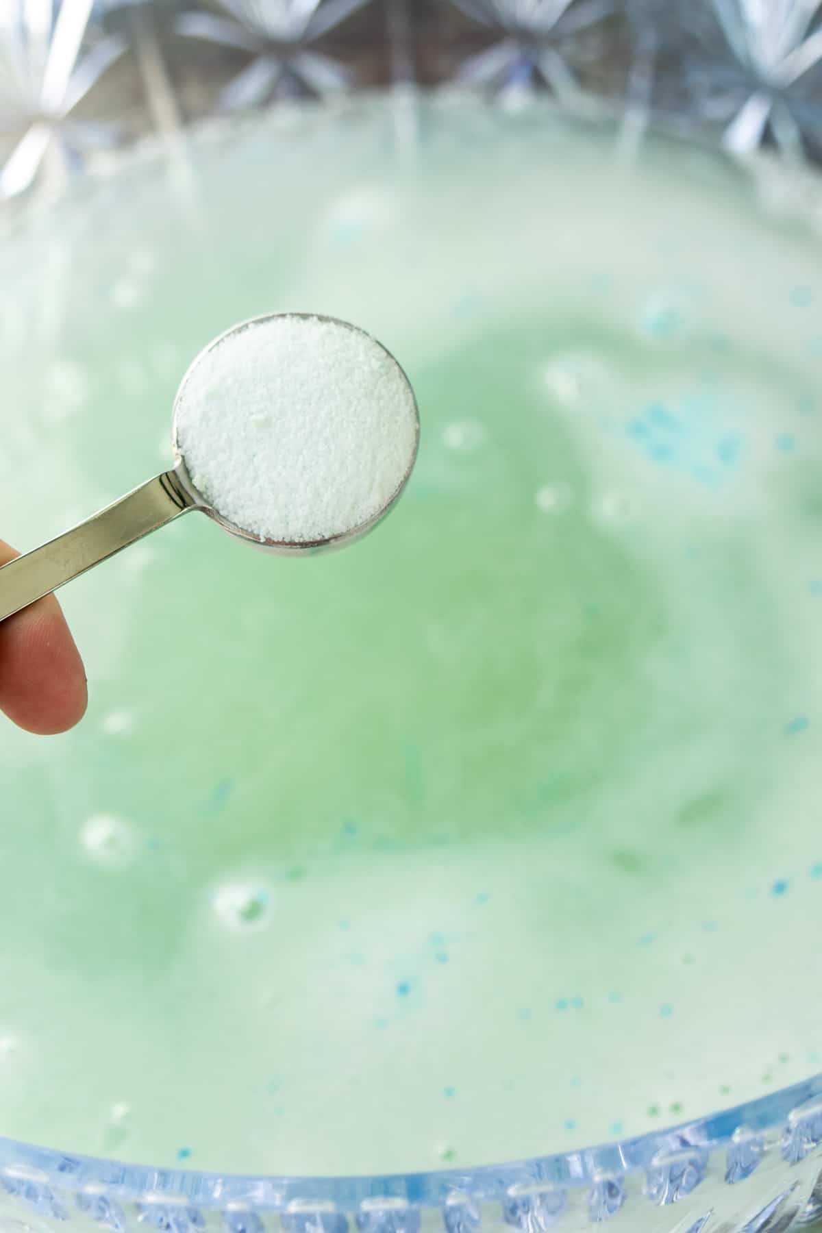 Hand holding a metal measuring spoon above a bowl of punch