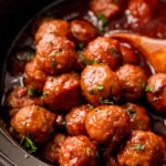 A crock pot full of grape jelly meatballs with a wooden spoon