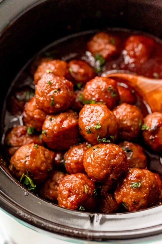 A crock pot full of grape jelly meatballs with a wooden spoon