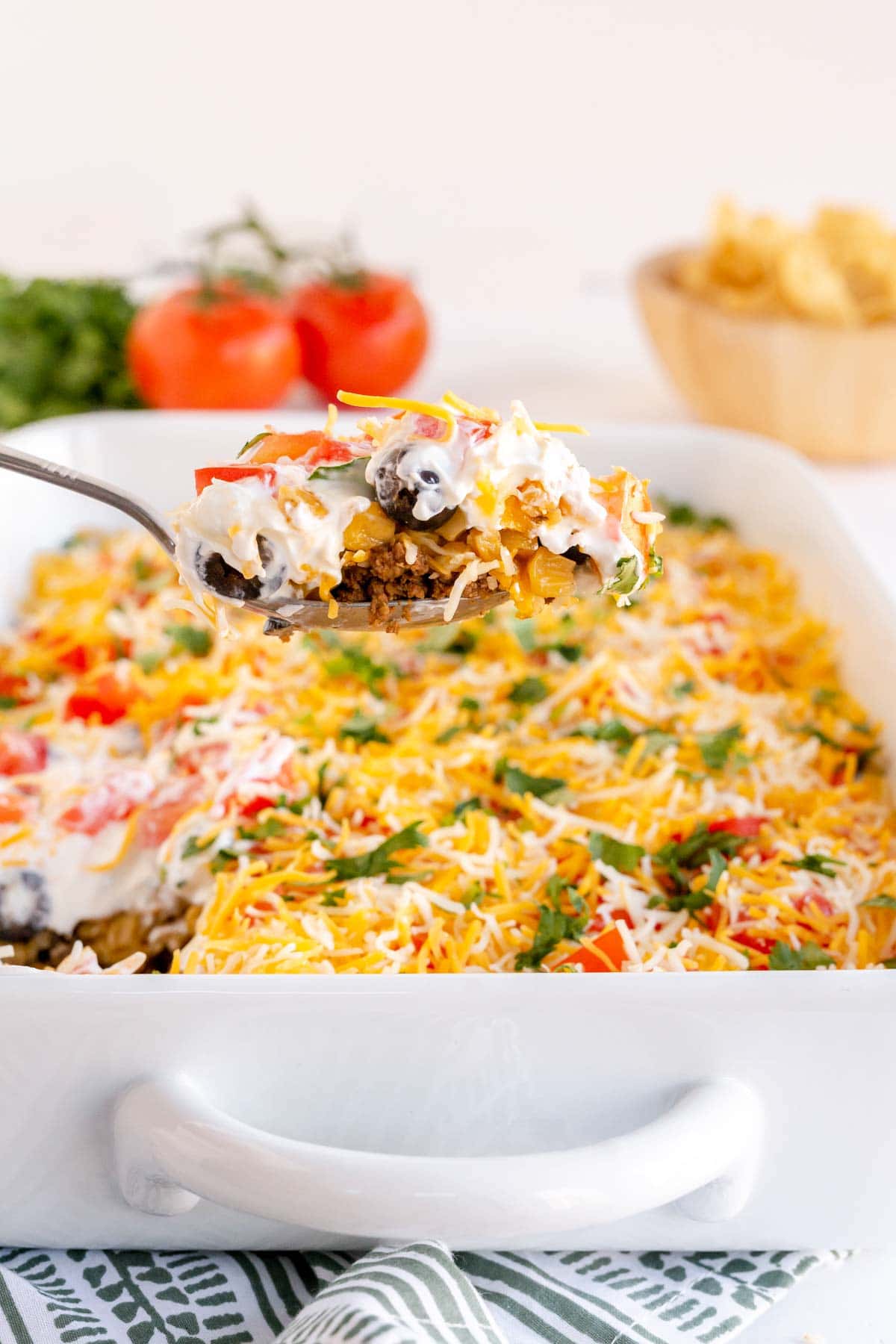 A spoon full of layered taco dip above a baking dish full