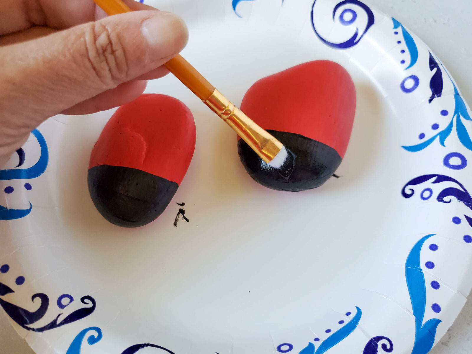 woman's hand painting a black face on a ladybug rock