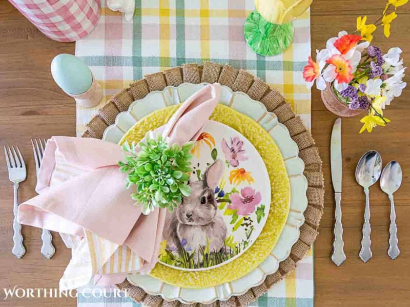 gingham table runner with multicolored plates stacked and topped with a pink napkin and floral napkin ring